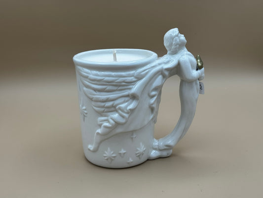 White ceramic mug candle with angel design on handle against a beige background